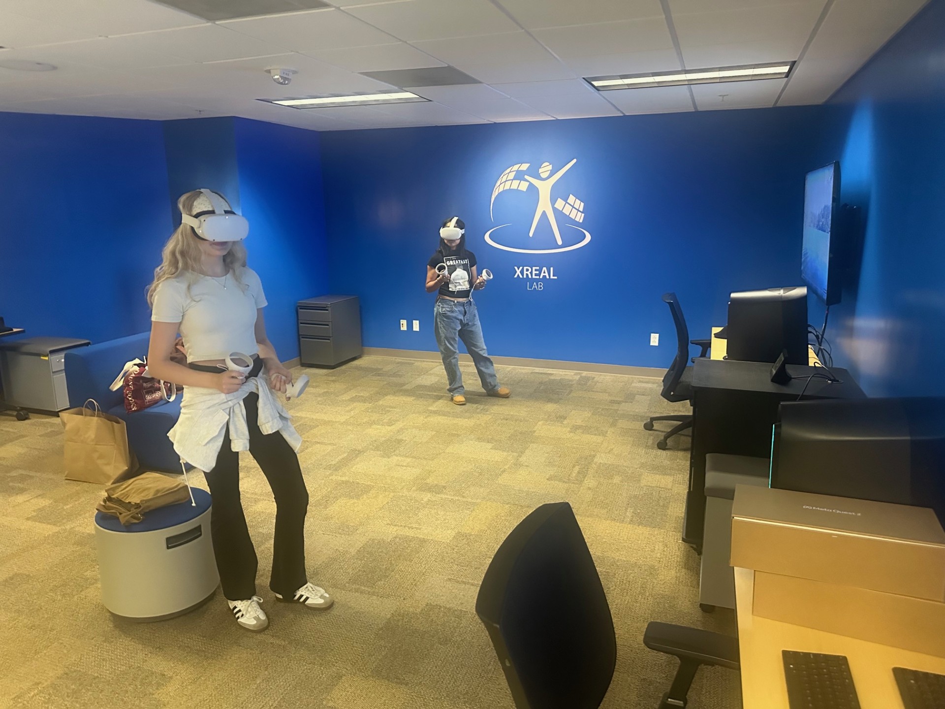 Two people wearing **Meta Quest 2 VR headsets** explore virtual experiences inside a bright blue room labeled **xREAL Lab**. The space includes computers, chairs, and open floor area for movement, creating a dedicated environment for immersive learning and training.