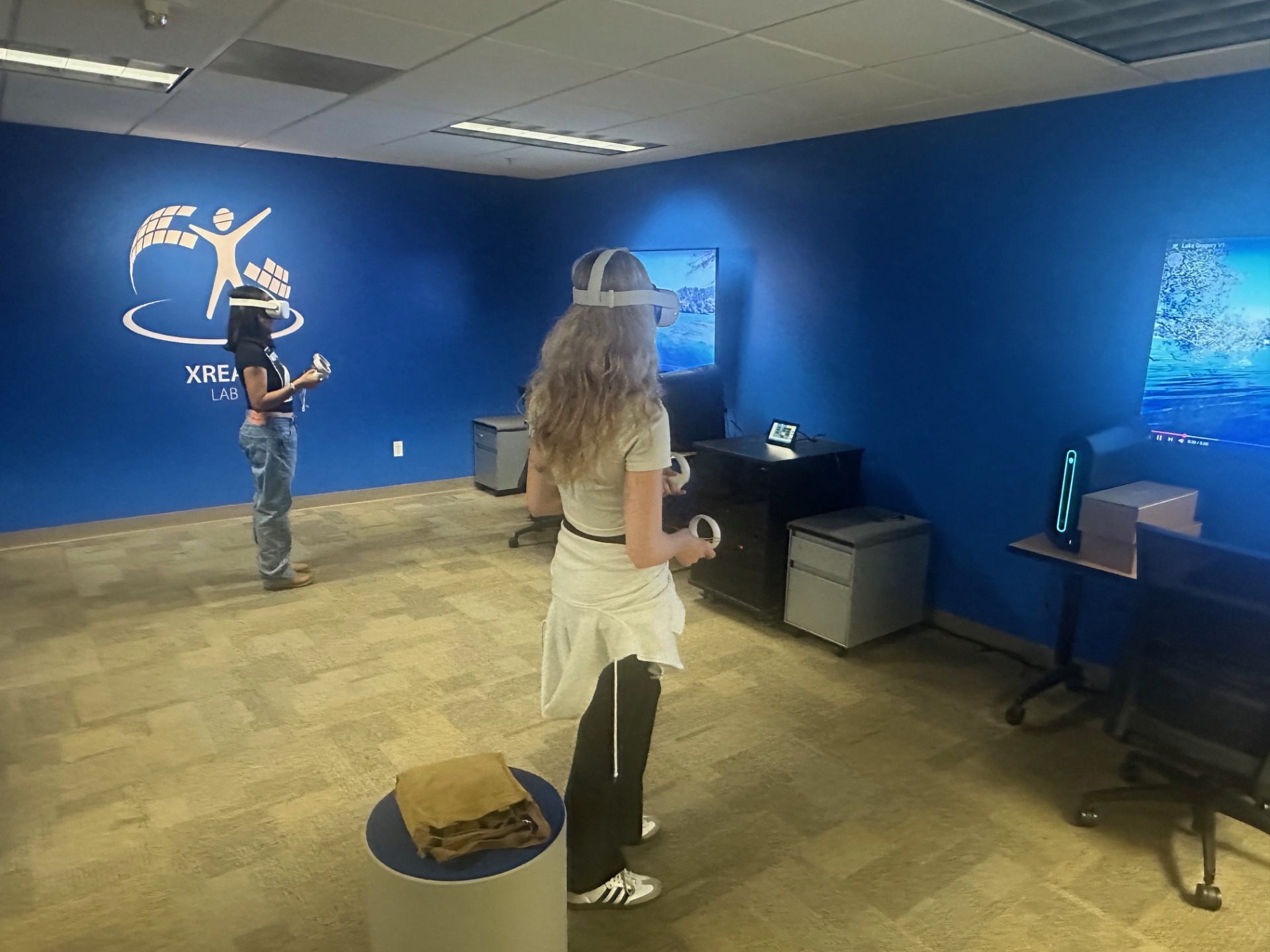 Two people wearing virtual reality headsets and holding controllers in a blue-walled room labeled “xREAL Lab.” One person