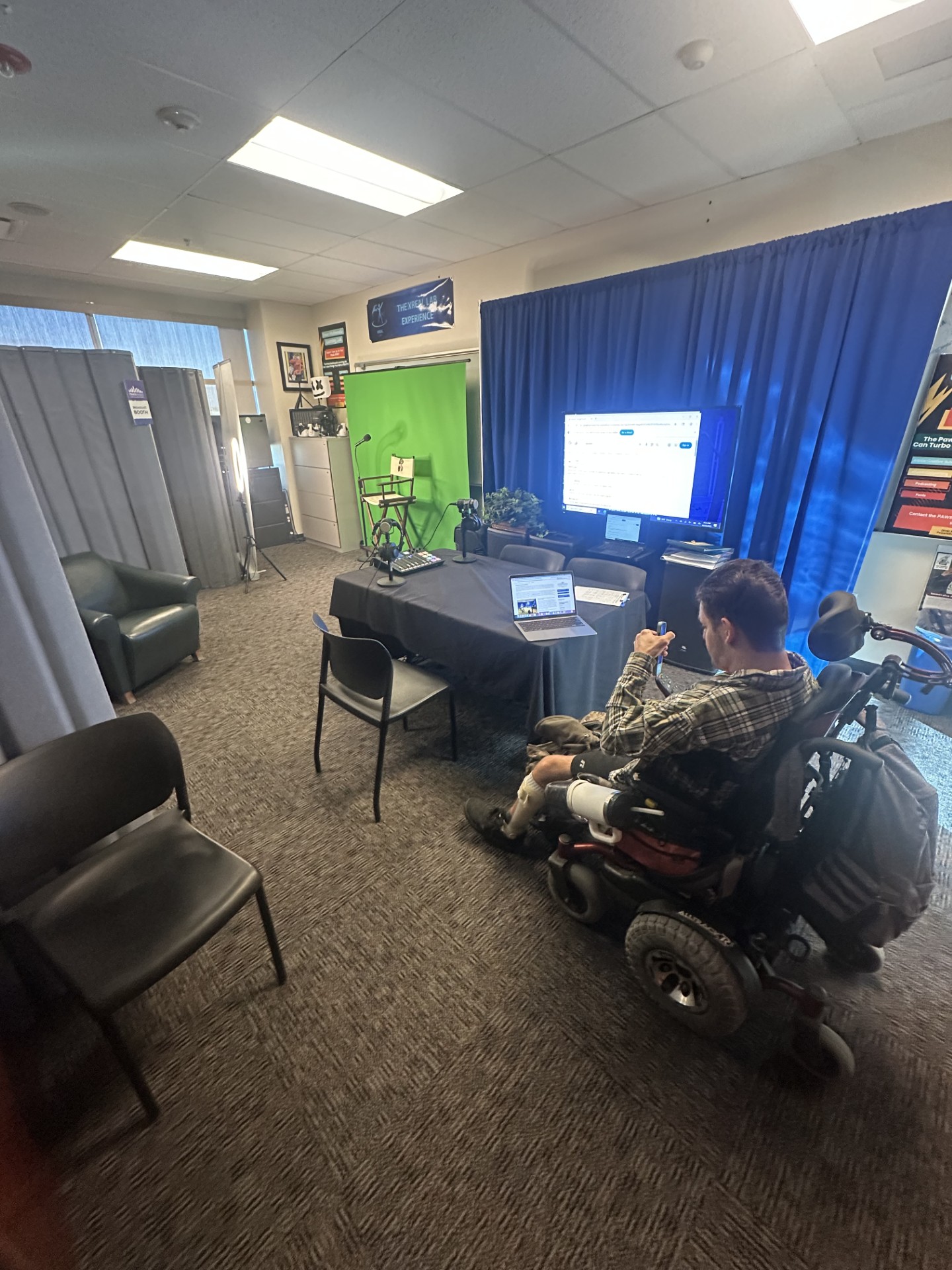 A multimedia recording studio with a green screen, lighting equipment, microphones, a large monitor, and a table set up for production. A person in a motorized wheelchair is working at a laptop in the foreground. The room has blue curtains, posters, and professional audio-visual gear.