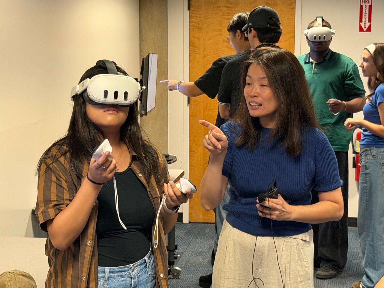 An instructor guides a student wearing a virtual reality headset and holding two controllers while other students participate in VR activities in the background.