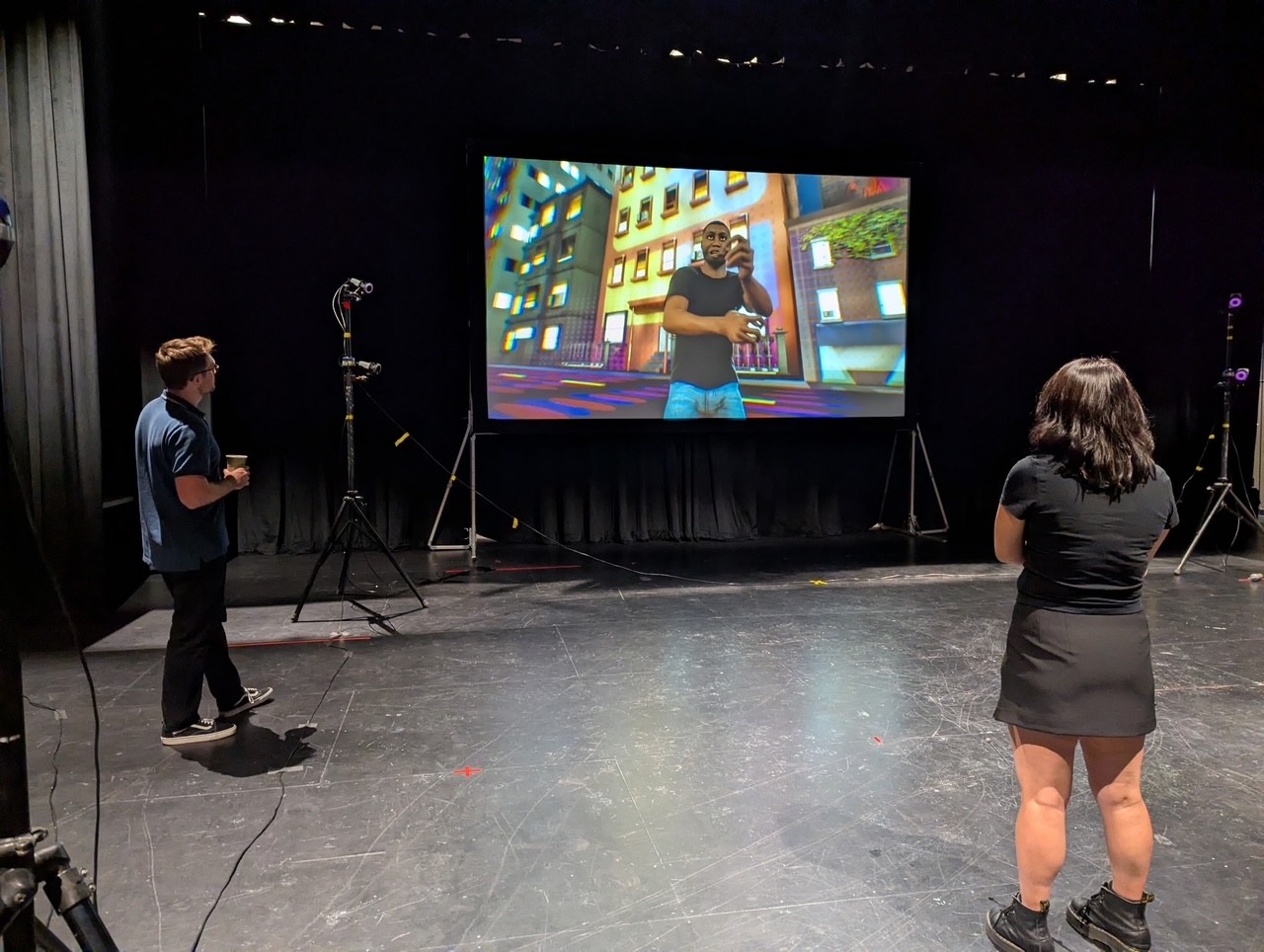 Two people stand on a stage facing a large projection screen that displays a 3D-rendered man in a city environment. The stage is set up with motion capture equipment, including cameras on tripods and tracking devices. The surrounding space is dark with black curtains, and the individuals appear to be observing or reviewing the virtual scene.