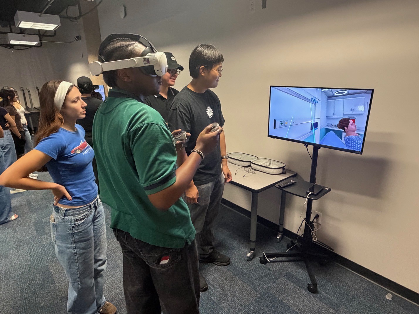 A student wearing a virtual reality headset uses handheld controllers to navigate a healthcare simulation while classmates watch on a monitor.