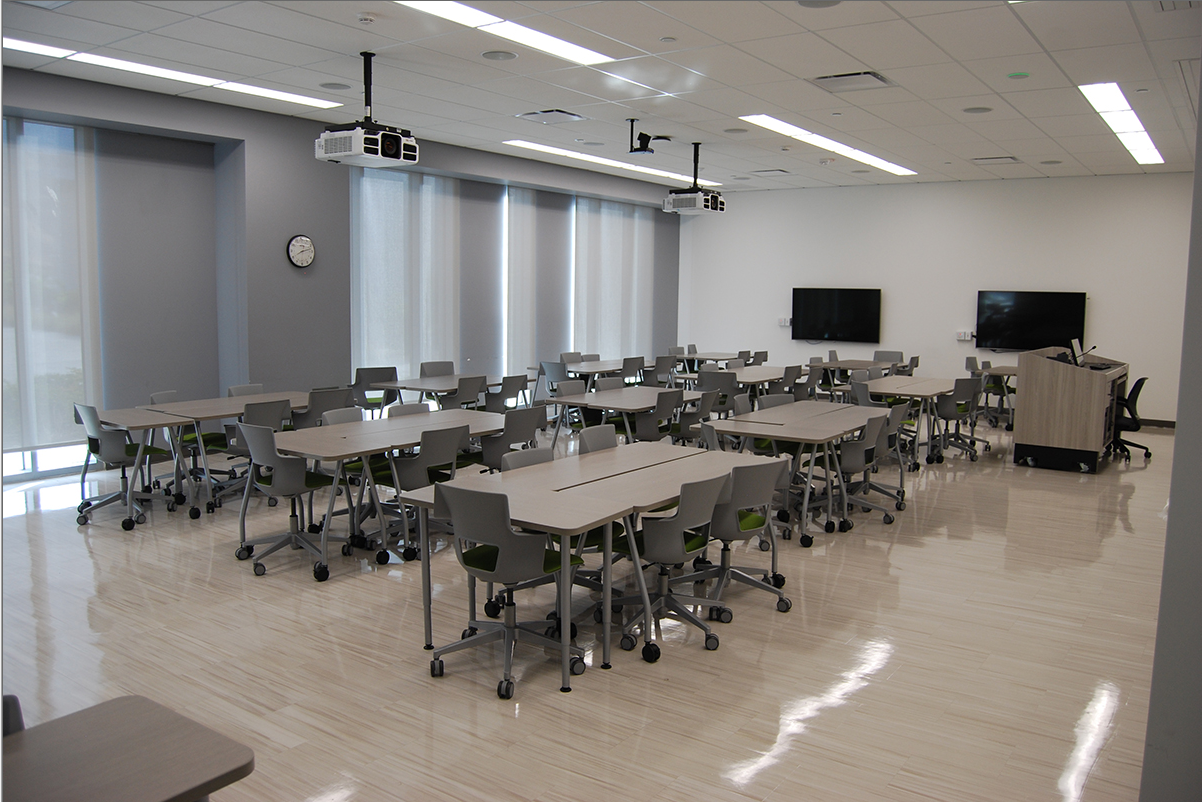 Modern active learning classroom with grouped tables and rolling chairs, ceiling-mounted projectors, wall-mounted displays, and large windows with shades letting in diffused light.