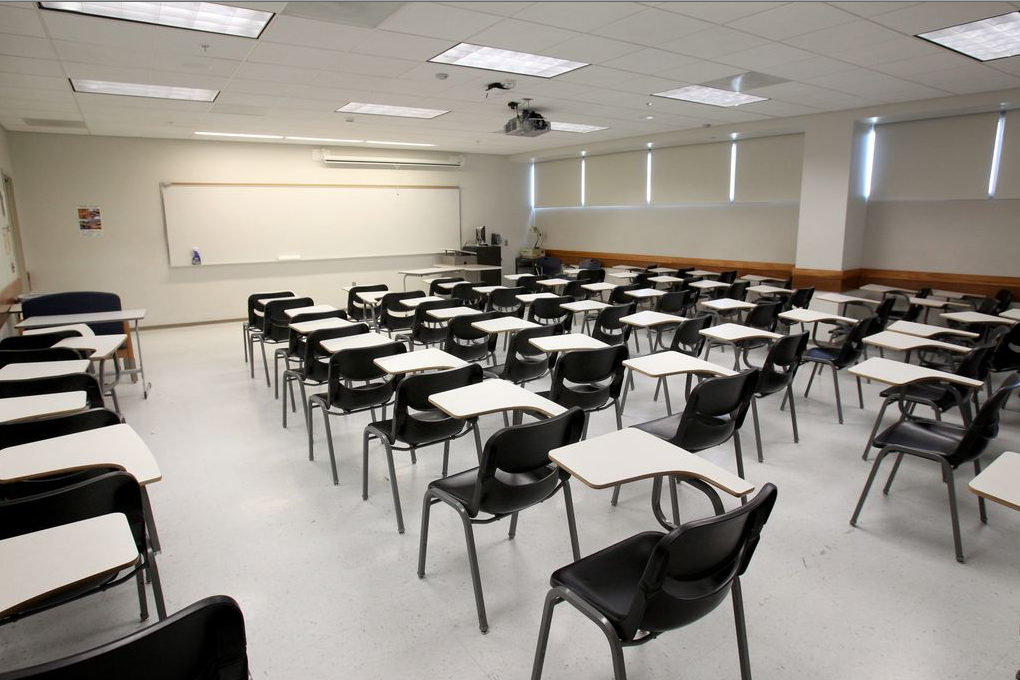 Bright classroom with rows of individual desk-chair combinations facing a large whiteboard, a ceiling-mounted projector, and windows with partially closed blinds along one wall.