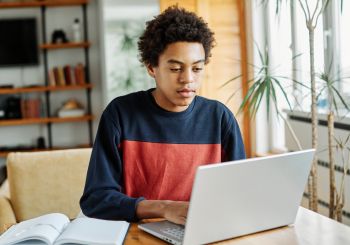 Student typing on a laptop with an open book to the side.