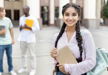 Student standing outside with a book in her hands and a light green backpack.She has long black hair in braids that come to the front.