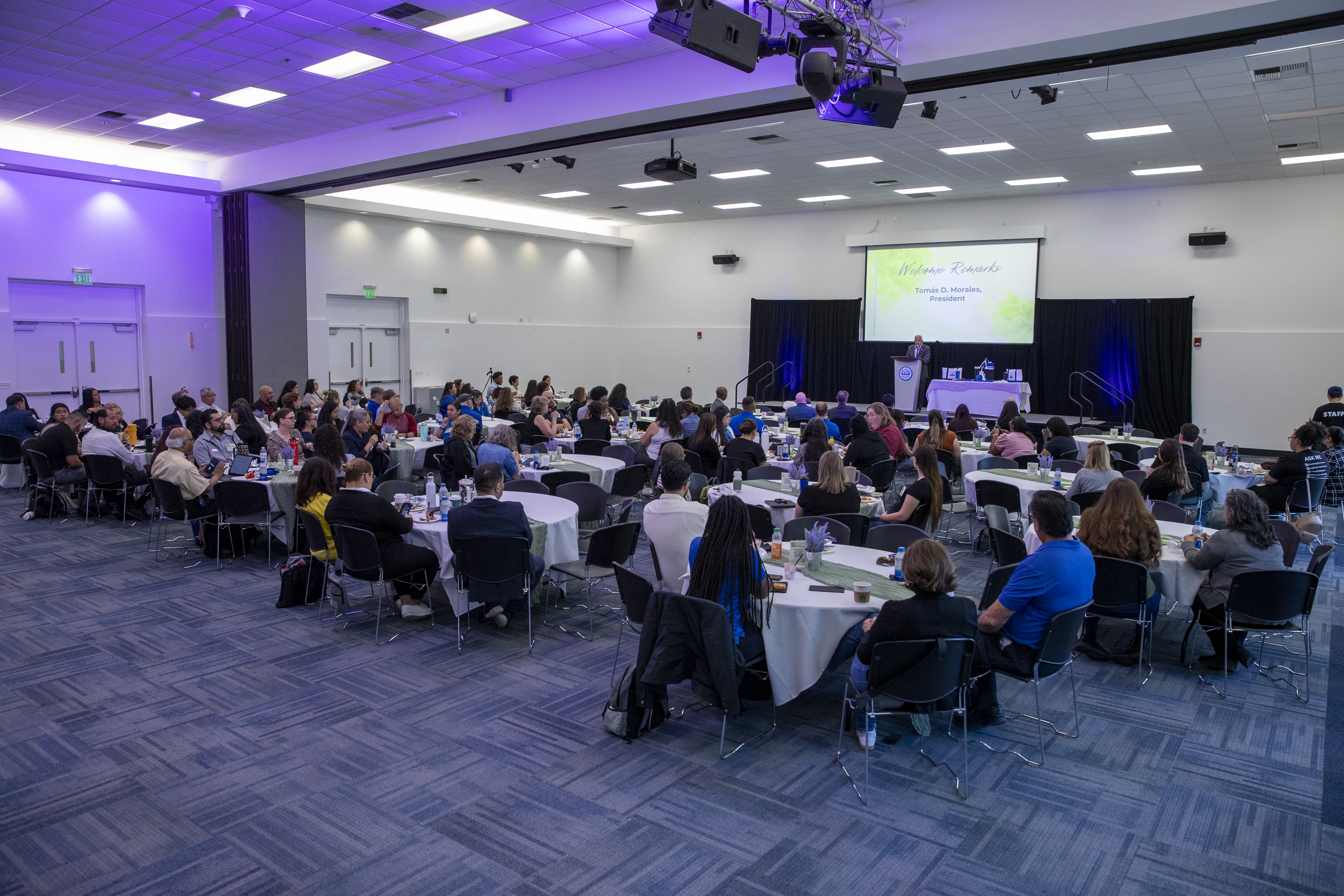 Wide-angle view of a 2026 CSUSB Business Conference with attendees seated at round tables facing a stage where a speaker delivers welcome remarks under a projected screen.