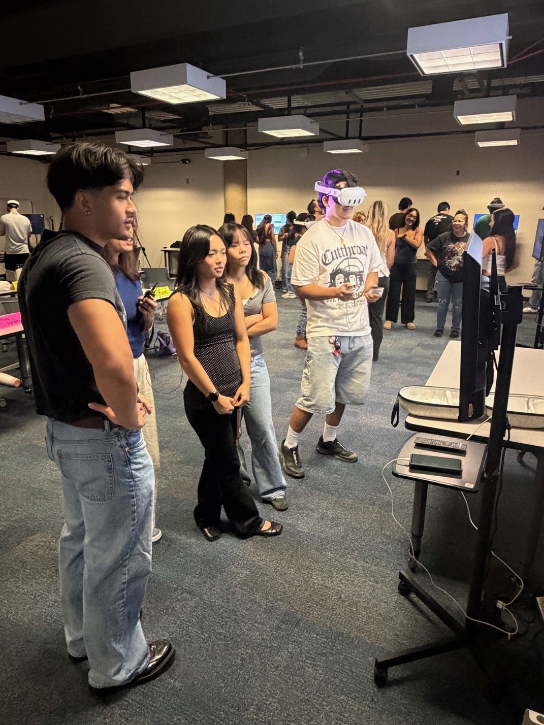 A student wearing a virtual reality headset uses two controllers while classmates observe the experience on a nearby monitor in a busy immersive learning classroom.
