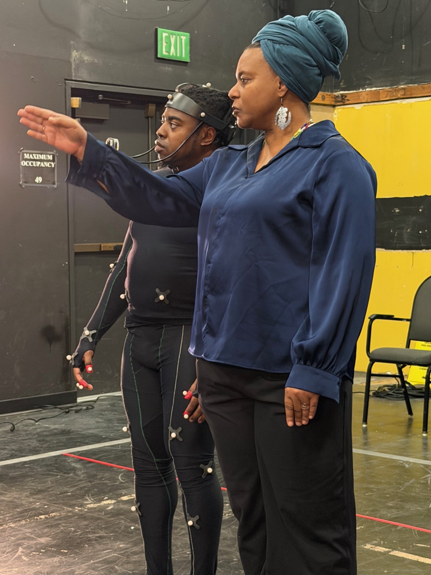 A woman in a blue blouse and headwrap stands on a stage, extending her arm and pointing forward while looking in that direction. Behind her, another person wearing a black motion capture suit with tracking markers and a head-mounted device looks the same way. The setting appears to be a theater or studio space, with a visible exit sign, a maximum occupancy sign, and stage flooring.