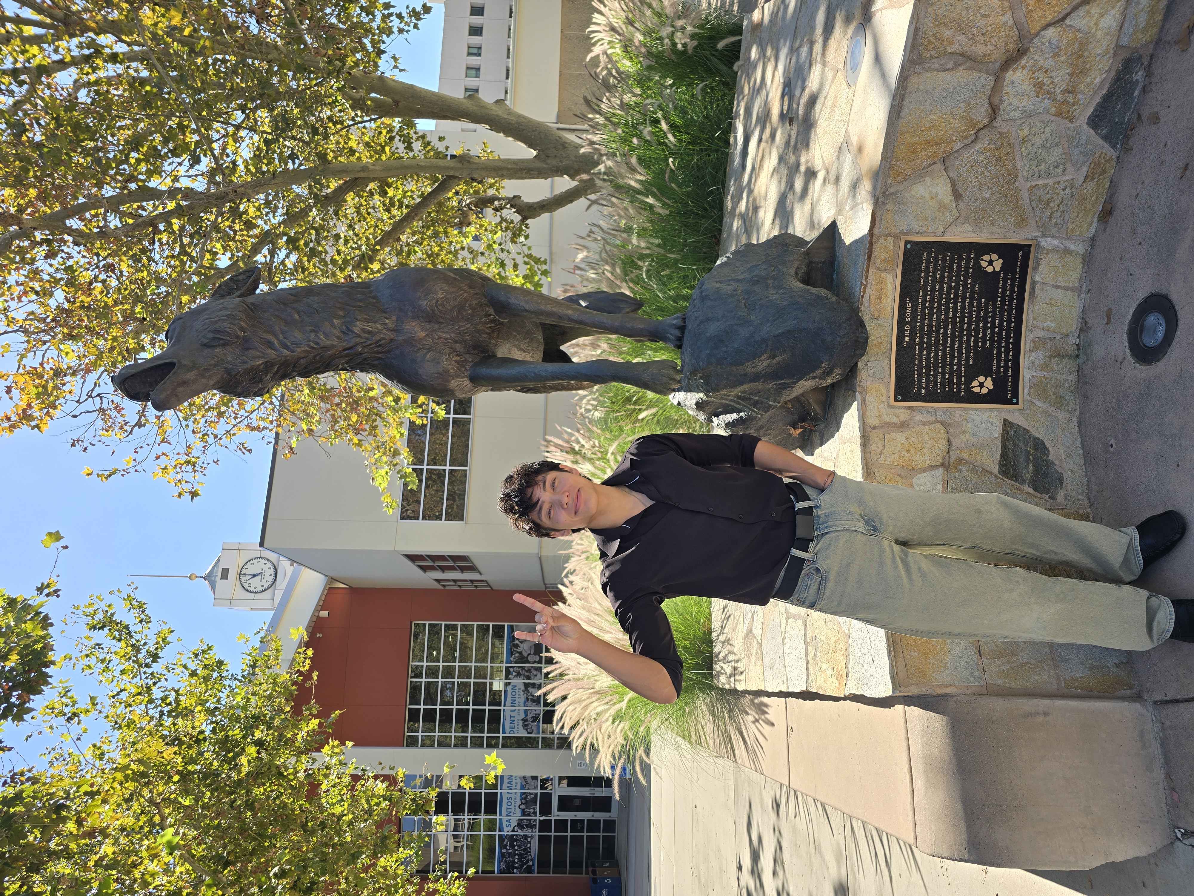 A male student in a black shirt and khaki pants making a Coyote hand gesture beside a bronze howling coyote statue on a sunny, tree-lined campus.