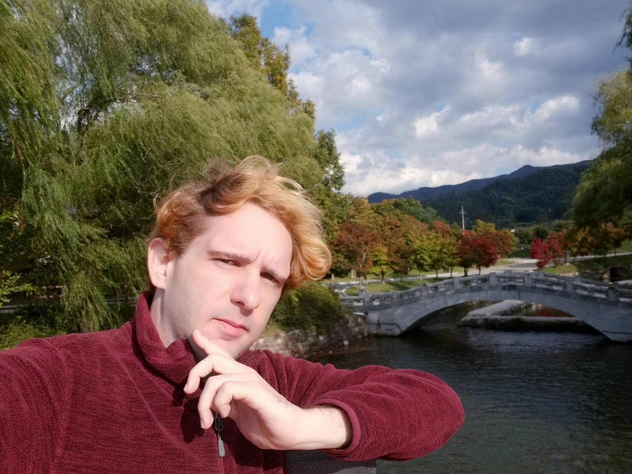 Cody Vrba, red sweater, stands in front of a bridge and landscape