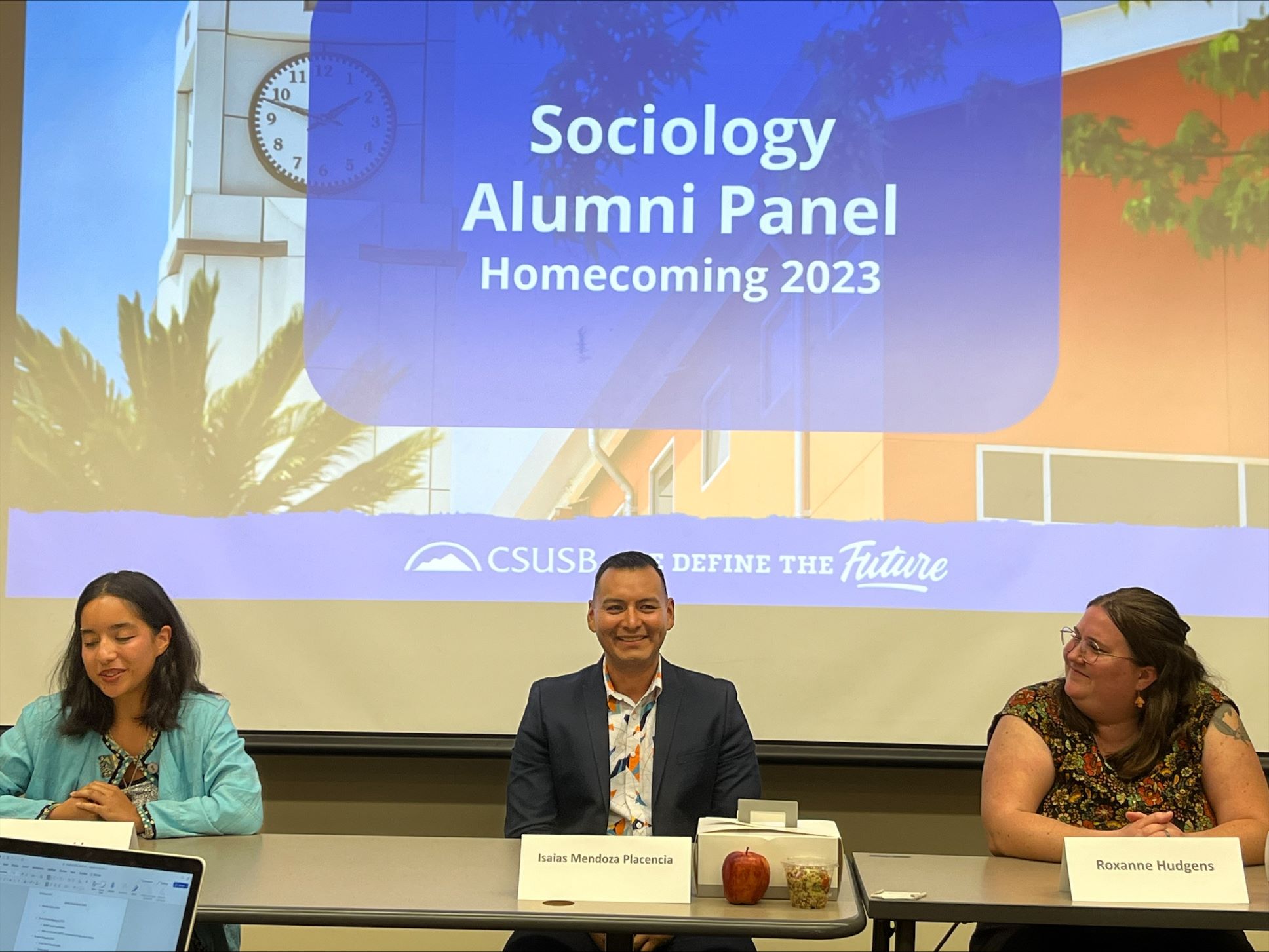 The image depicts three people sitting at a table facing an audience with the text "Sociology Alumni Panel Homecoming 2023" behind them.
