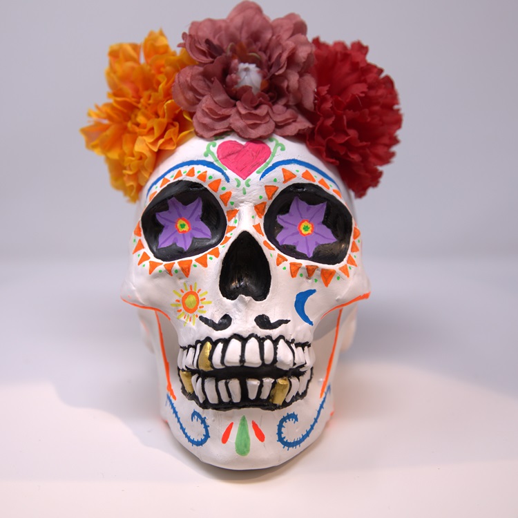 White painted in a sugar skull style with orange, blue, pink and black details, and a crown of flowers.