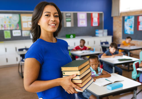 African American female elementary teacher smiling and holding books with students in the background