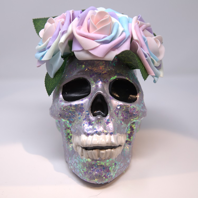 Metallic iridescent painted skull which glows in the dark, adorned with a flower crown and glitter.