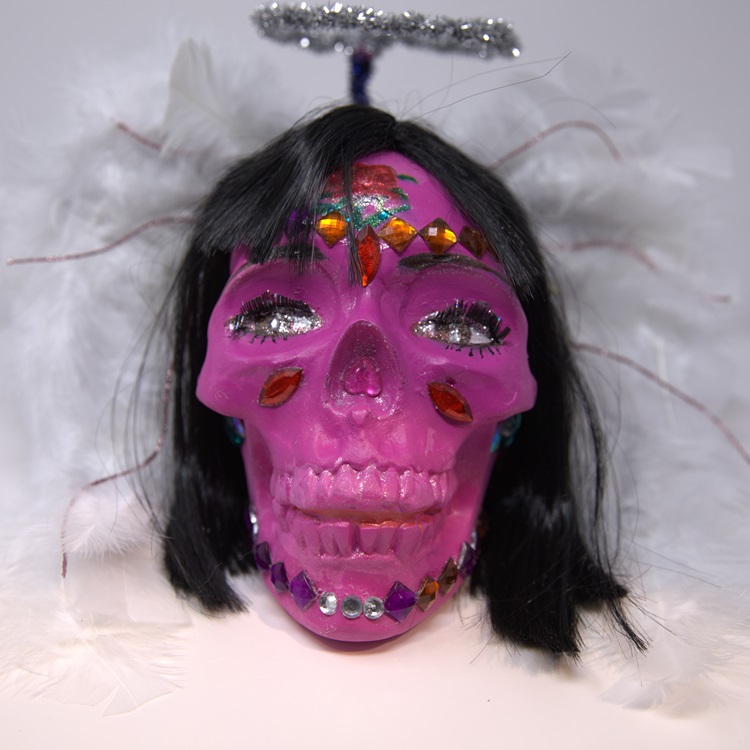 Pink painted skull adorned with gemstones, it's features include black hair and lashes, angel wings, and a silver halo.