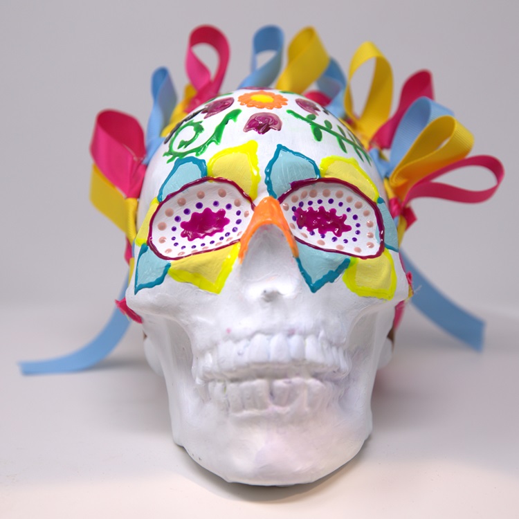 White skull with decorative flower and leaf patterns in yellow, turquoise, magenta and orange, adorned with a crown of ribbons.
