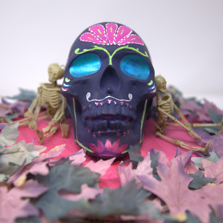 Purple painted skull with flower details in green and metallic pink and metallic blue eyes, surrounded by leaves and mini skeletons.