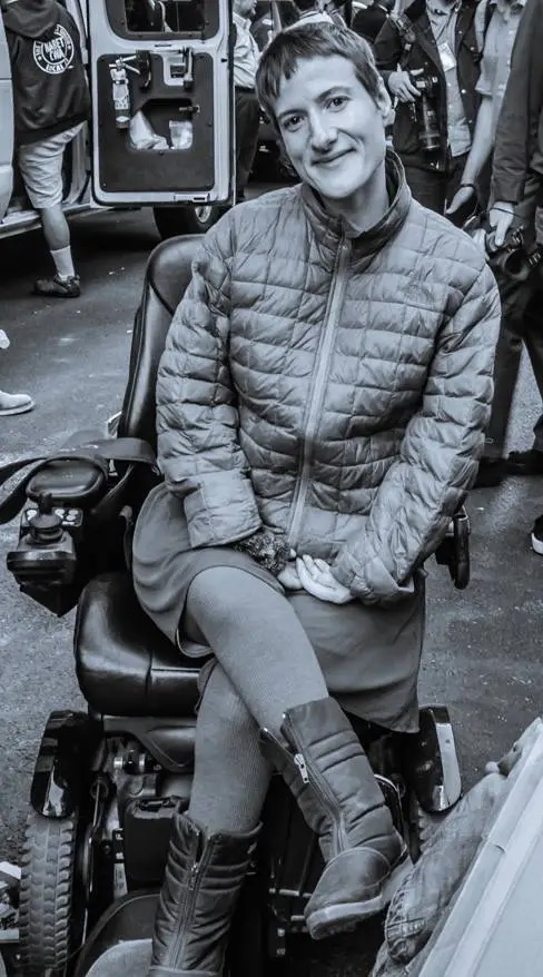 Sunaura Taylor, black and white image, woman with short hair in puffy jacket sitting and smiling in wheelchair