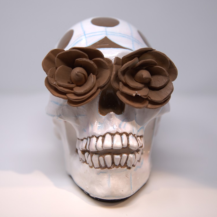White skull with brown dots, blue linear patterns, and hand crafted clay flowers on the eyes.